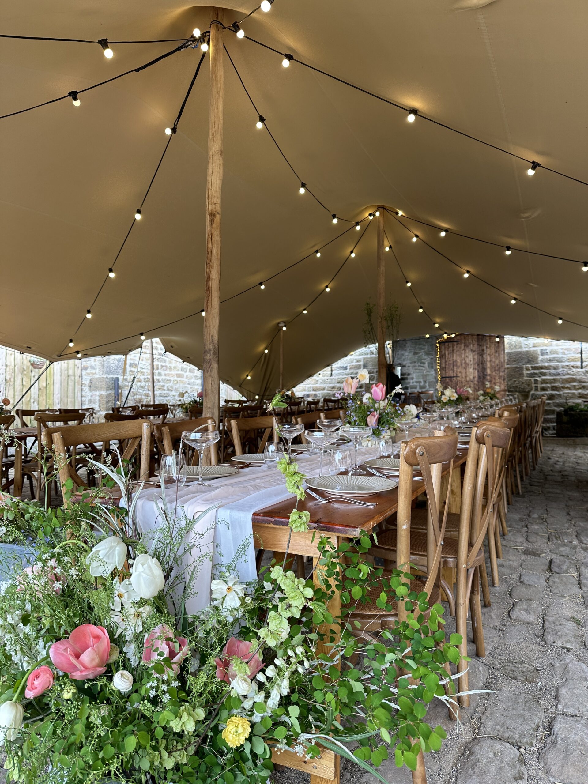 stretch tent by Countryside Events with festoon lighting and rustic dining tables and spring flowers by Forge Events, situated in the courtyard at The Owl Barn, Little Seed Field, North Yorkshire