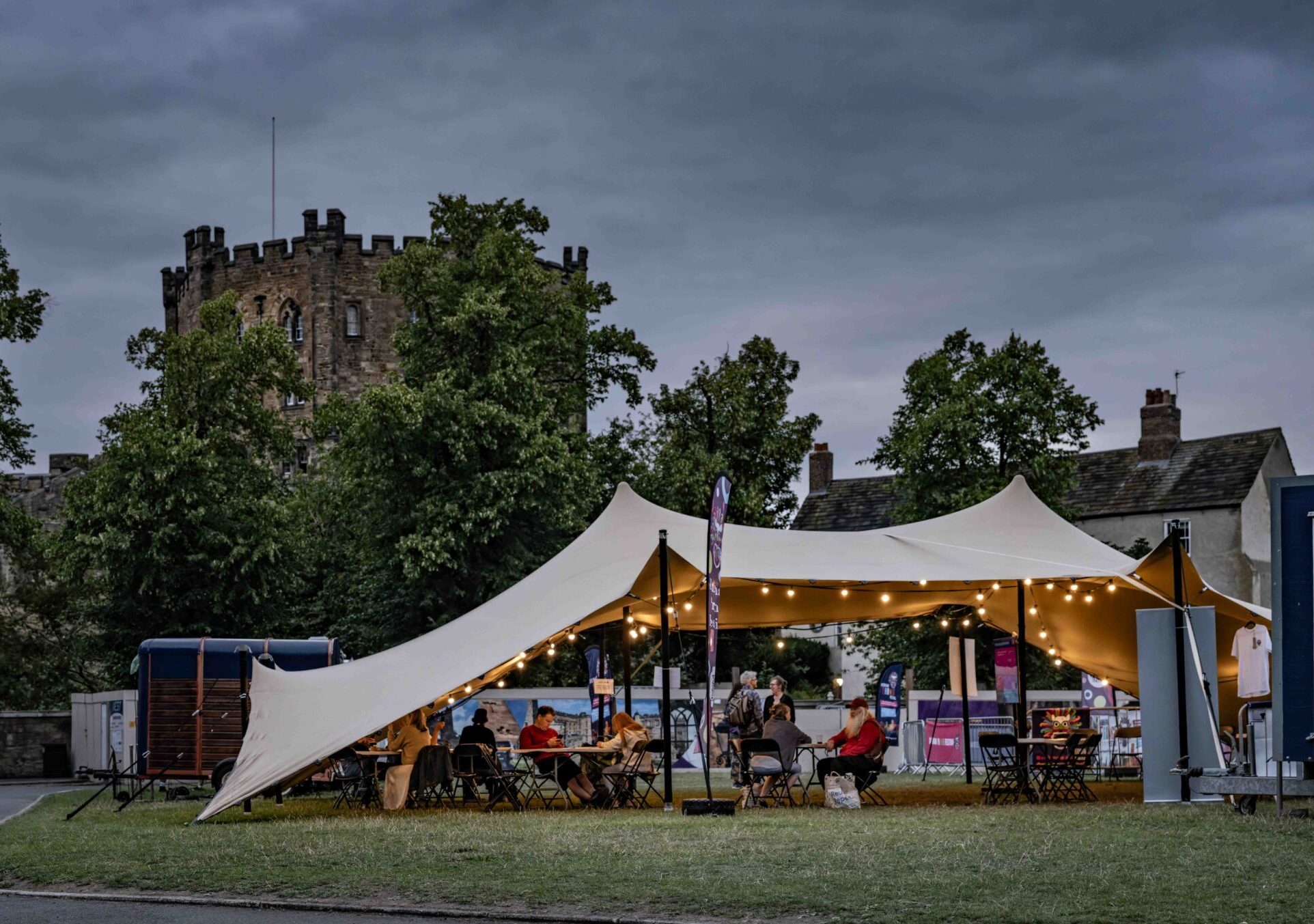 a stretch tent at dusk, Durham Fringe, with festoon lights and visitors sat underneath canvas